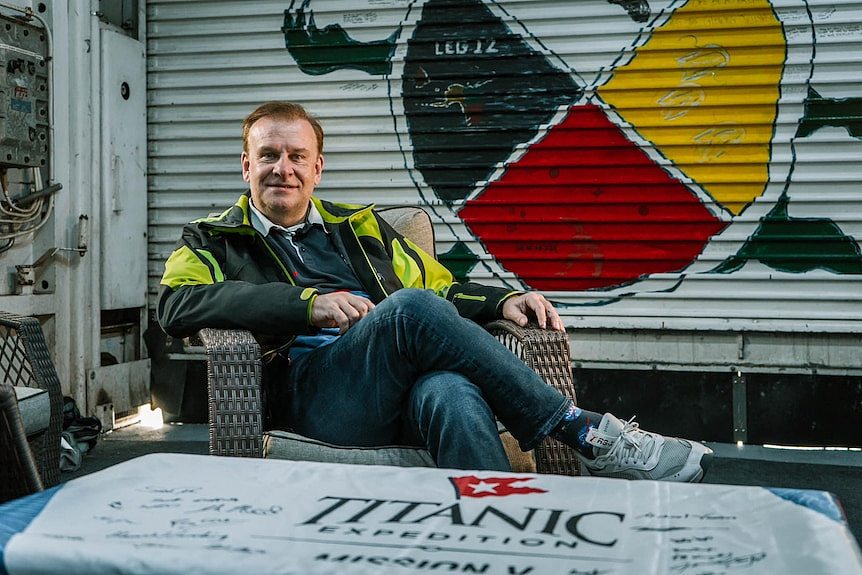 A middle-aged man in jeans and a jacket sits in a chair and smiles. In front is a table with Titanic Expedition written on it