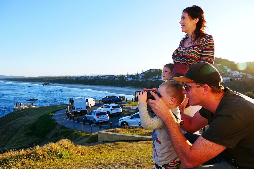Father holding binoculars up to child's face pointing out to sea