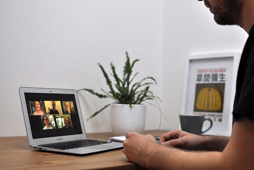 A man sits at a table participating in a Zoom meeting. On the laptop in front of him are five faces of people in the meeting.