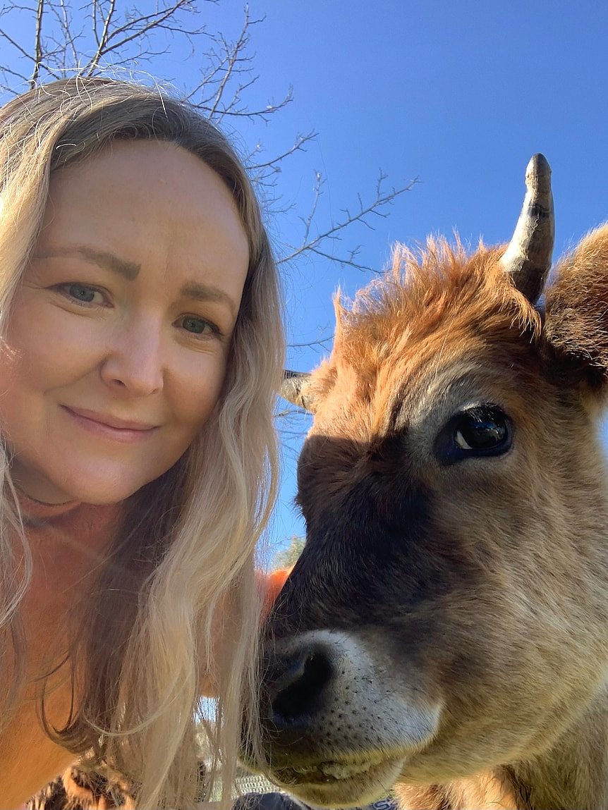 A woman with long blonde hair smiles next to a cow's face.