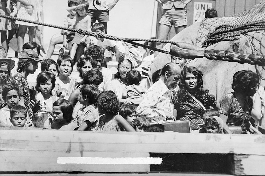 Black and white photo of large crowd of people, many squinting into the sun, aboard a boat, just visible.