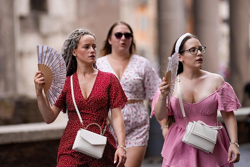 Three women in bright coloured dresses walk along Romes streets with foldable fans