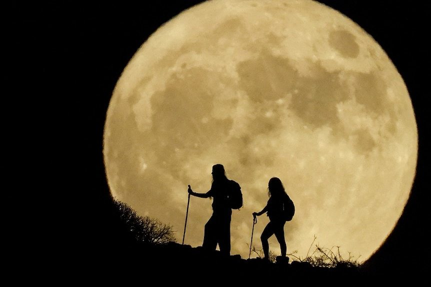 Two women hiking up a hill with a full, yellow moon behind them
