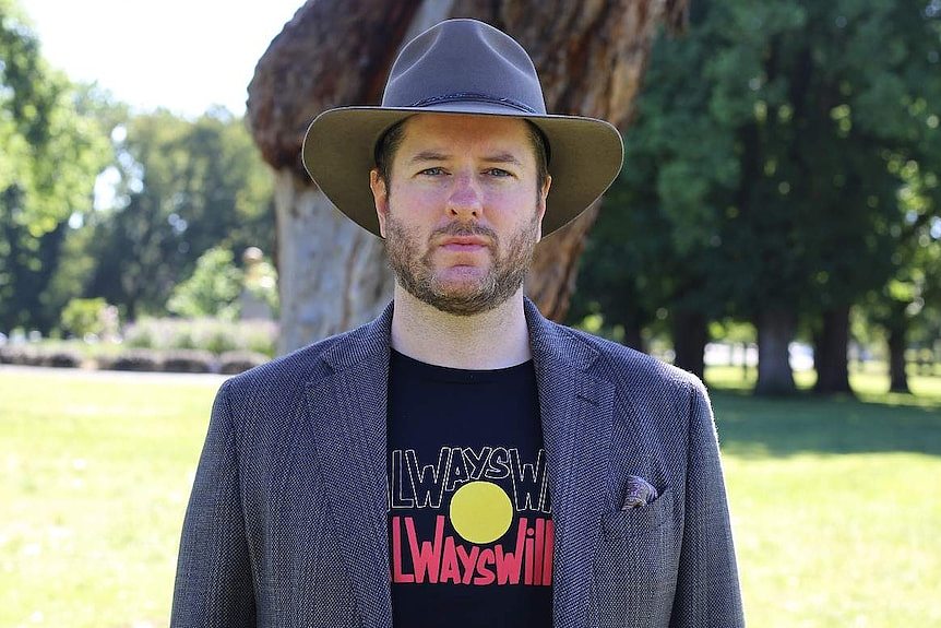 Marcus Stewart stands in a park, dressed in an Akubra and t-shirt bearing the message 'Always Was, Always Will Be'.