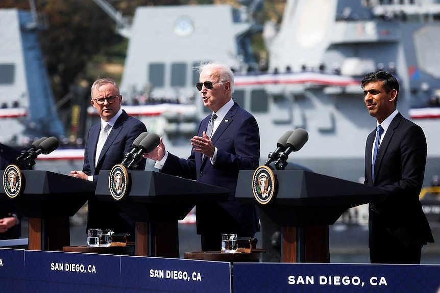 Anthony Albanese (left), Joe Biden (middle) and Rishi Sunak (right) all stand at podiums in front of ships.