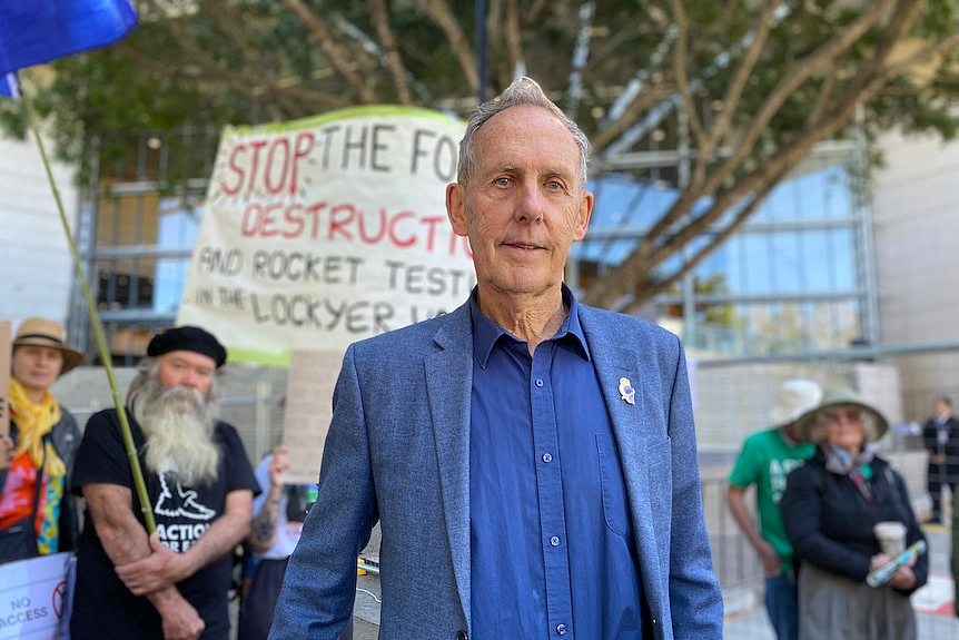 bob brown in a blue shirt and jacket with protesters behind him