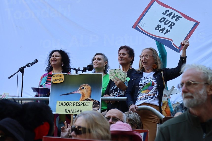 four women on stage behind mics holding signs saying save toondah