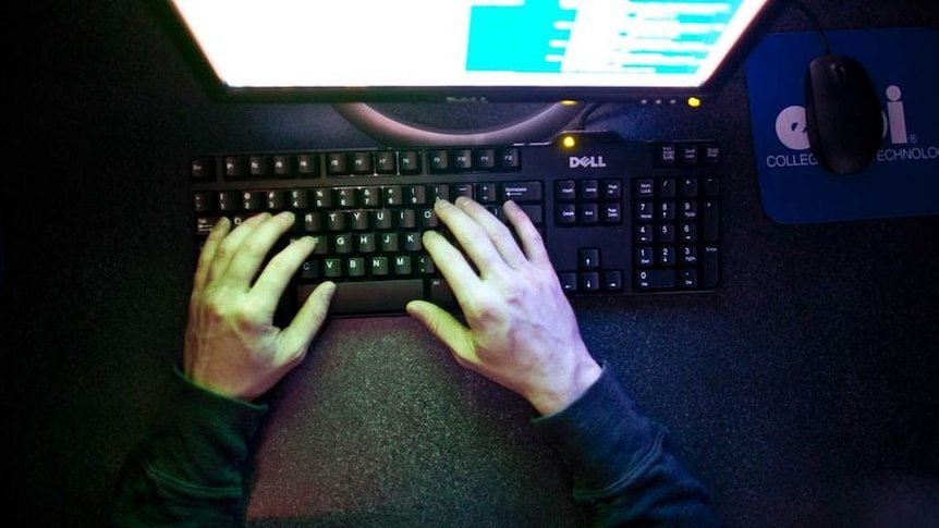 A person's hands are illuminated by the pale glow of a computer screen, as they type in a dark room.