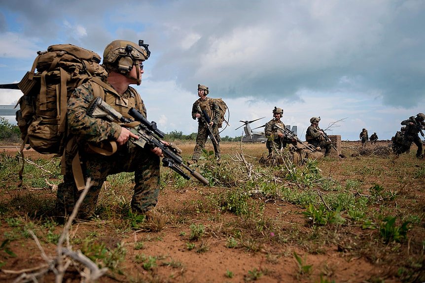 A small group of US Marines crouching or standing in a row in red dirt, holding rifles.