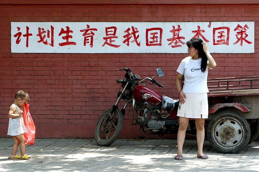 2002年，北京一位年轻的母亲在写有“计划生育是我国的一项基本国策”的标语牌前看着自己的孩子。