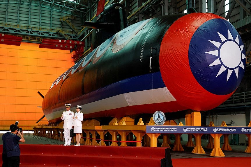 Two people stand in front of a large submarine with Taiwanese flag symbol draped over it.