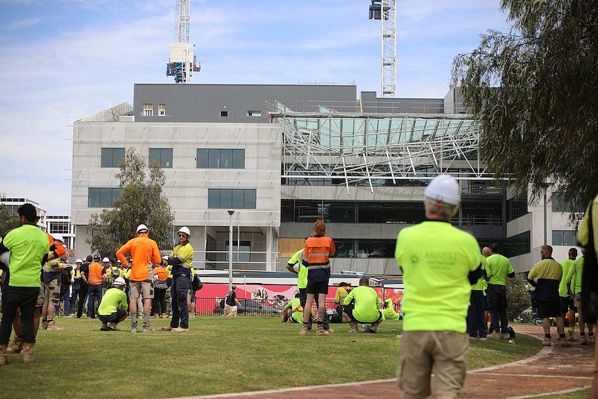 Workers in hi-vis clothing gathered on grass at Curtin University looking at the collapsed roof of a building.