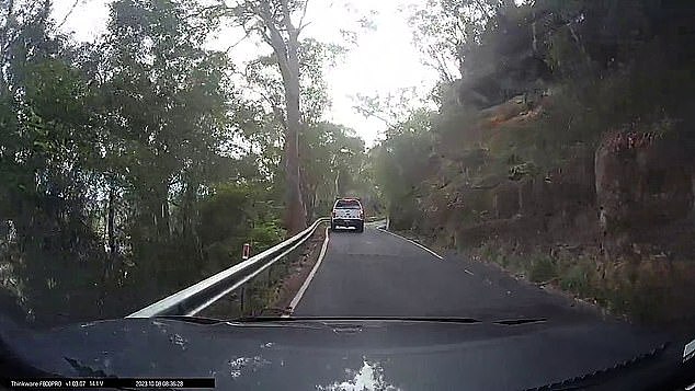 As the driver of a white Ford Ranger ute approaches a sharp curve going up a hill, a motorcyclist is suddenly seen emerging from the blind corner coming the other way