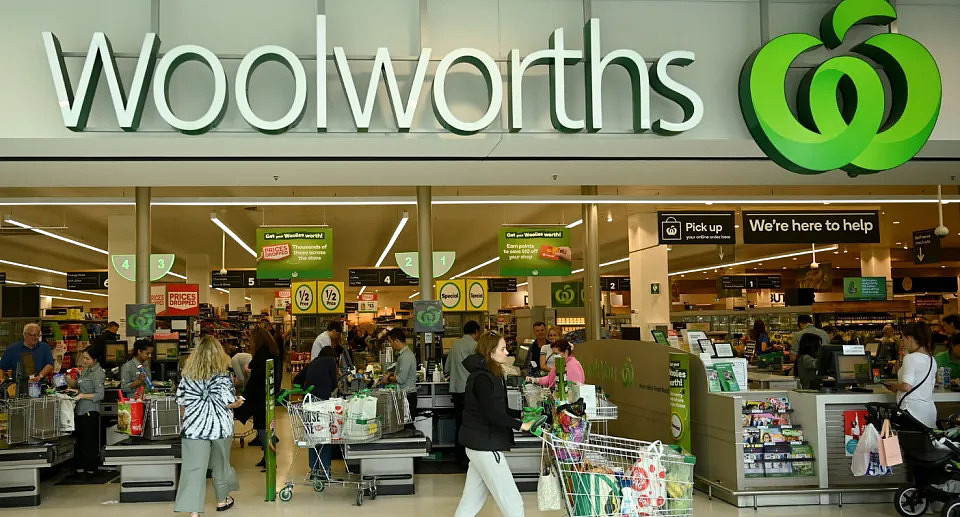 Photo out the front of a Woolworths store with the Woolworths sign at the top and shoppers pushing trolleys and checking out underneathe.
