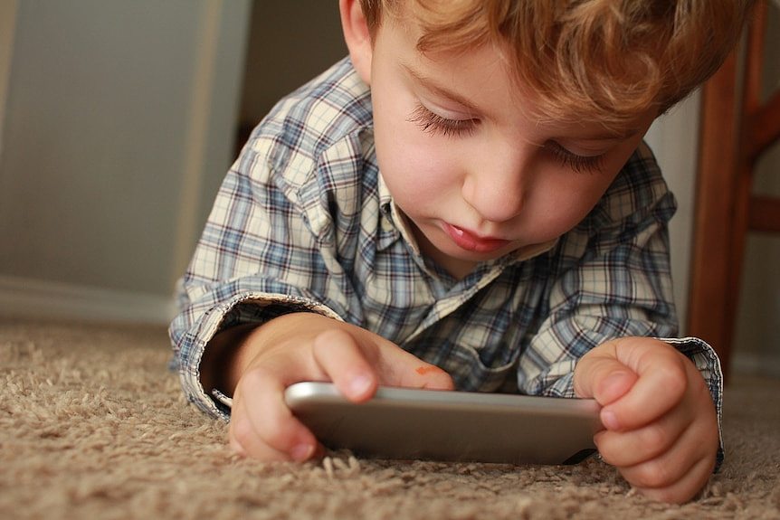 一个穿方格衬衣的孩子在地毯上玩手机。