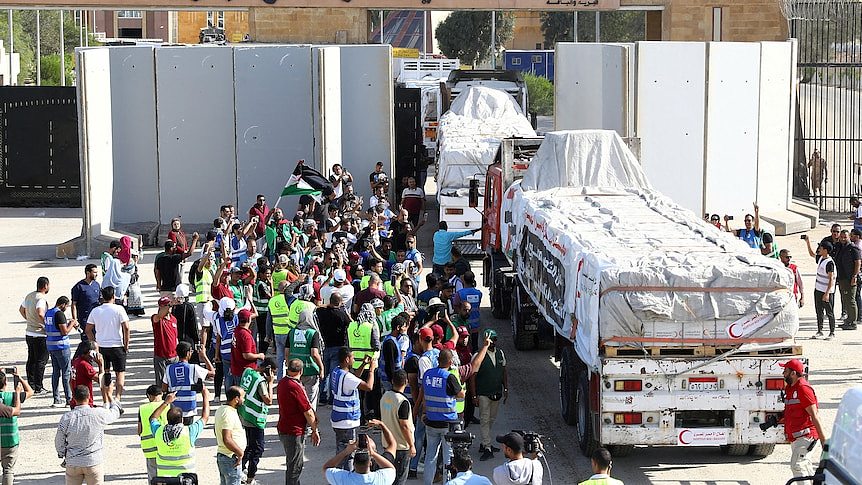 A crowd of people, some wearing hi-vis vests, gather beside several trucks loaded with covered goods as they emerge from a wall.