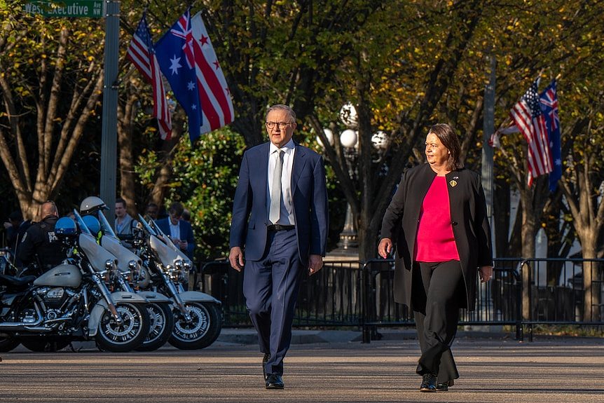 Anthony Albanese and Madeleine King walk past the White House. Australian and US flags are in the background.