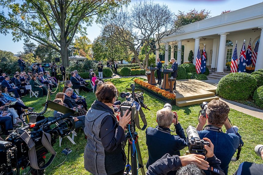 Reporters and photographers surround the two leaders, who are at podiums in front of Australian and US flags in a garden.