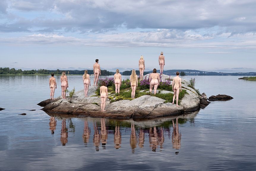 A photograph of 11 naked people standing on a rock, surrounded by water, facing away from the camera