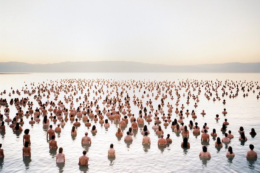 A large number of naked people standing in waist deep water, photographed from behind, looking at hills beyond