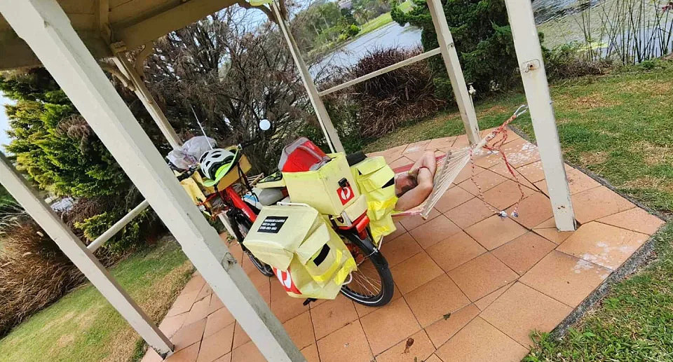 The Australia Post worker sleeps on his back with his arms above his head in the hammock with his bicycle parked to his left. 