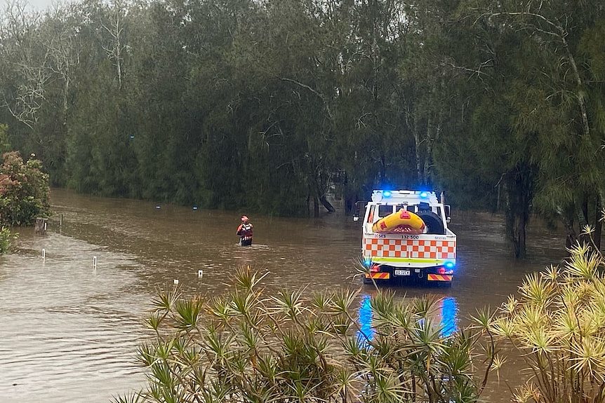 A person in high-vis wades through floodwater, near where an emergency vehicle is parked.