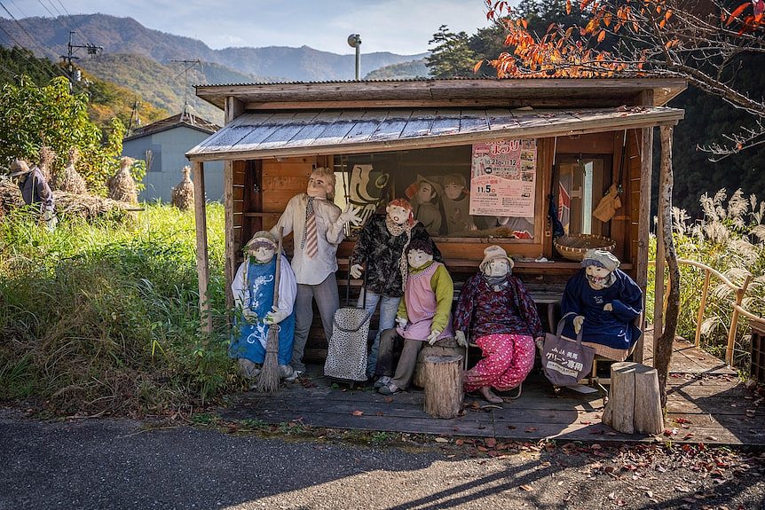 A group of six dolls sit gathered together at a bus stop.