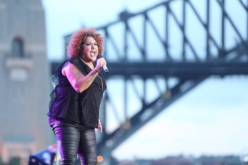 A woman with a black blouse and black leggings sings in front of the Sydney Harbour Bridge.