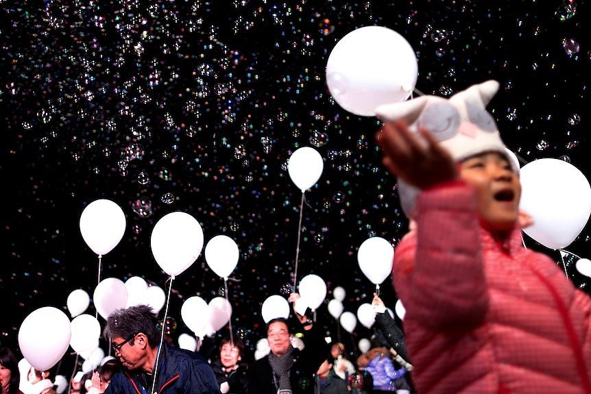 A group of people hold white balloons surrounded by bubbles in the night sky.