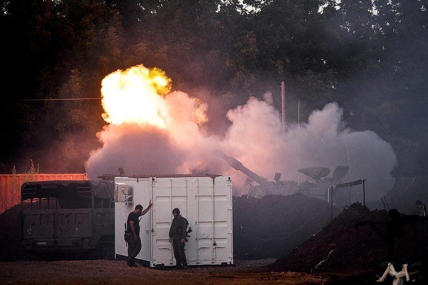 Two soldiers stand by a white shipping container as a large vehicle fires in a flash of smoke and flame behind them.
