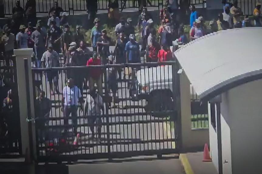 A crowd gathers outside a gate while several people try to break through the gate