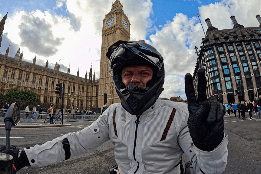 A man poses on a motorcycle in front of an old clock tower in a city