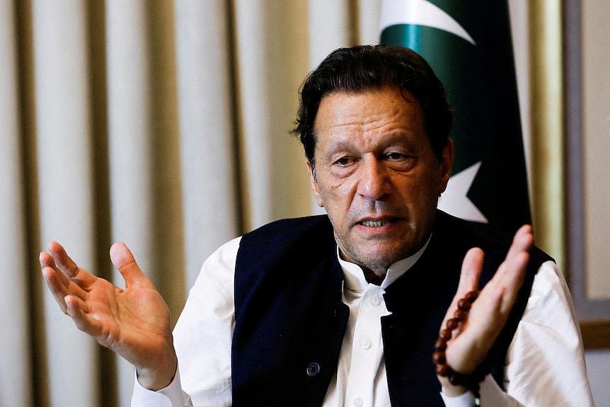 A close up of Imran Khan looking down and gesturing with his hands near a Pakistan flag.