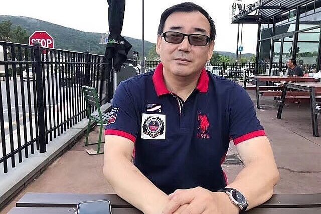 A photo of a Chinese-Australian man Yang Hengjun, sitting on an outside table, wearing a navy polo shirt with a red collar.