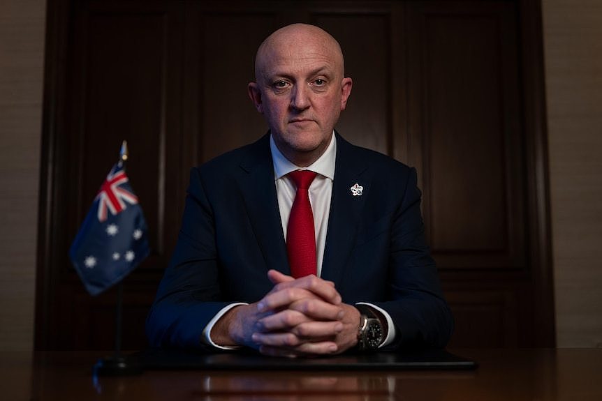 A bald man in a suit with his hands clasped on a desk 