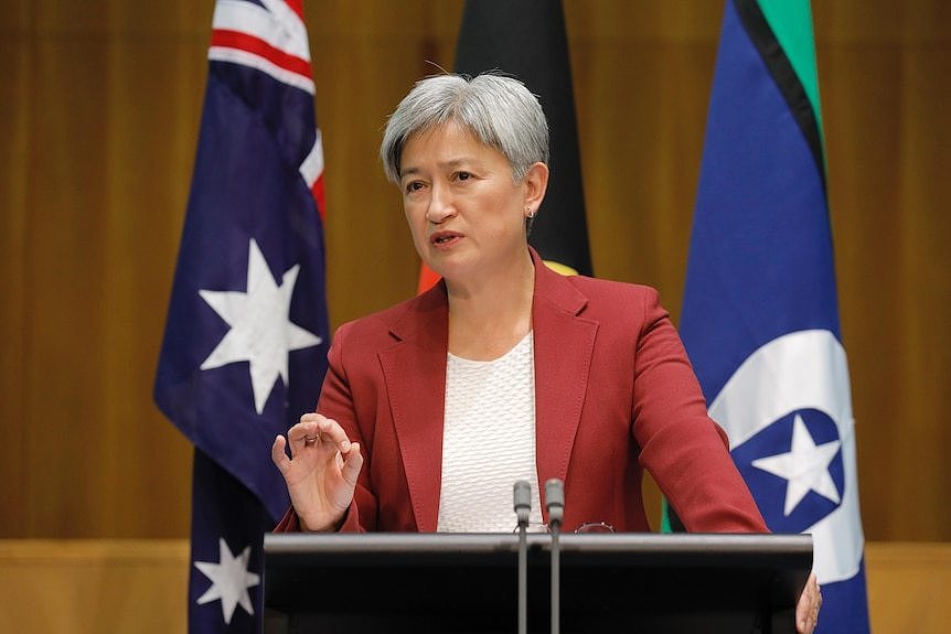 Penny Wong at a podium in a red blazer. 