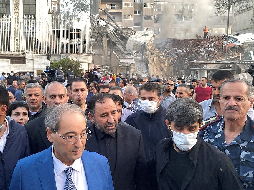 A man in a light blue suit leads a crowd of people walking away from a smouldering wrecked building