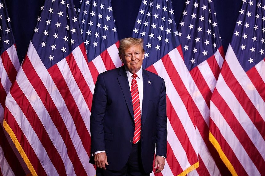 Donald Trump standing in front of a row of American flags smiling