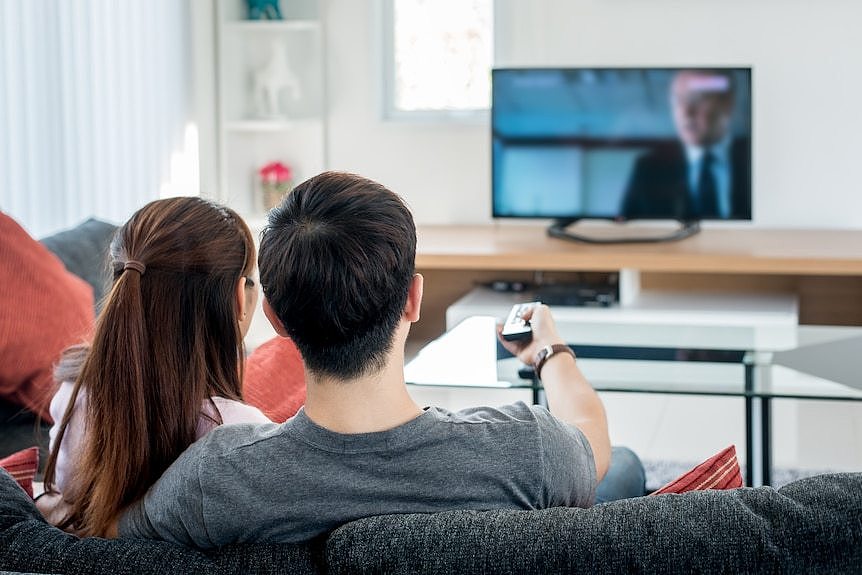 A couple sit watching TV, the man holds up a remote control