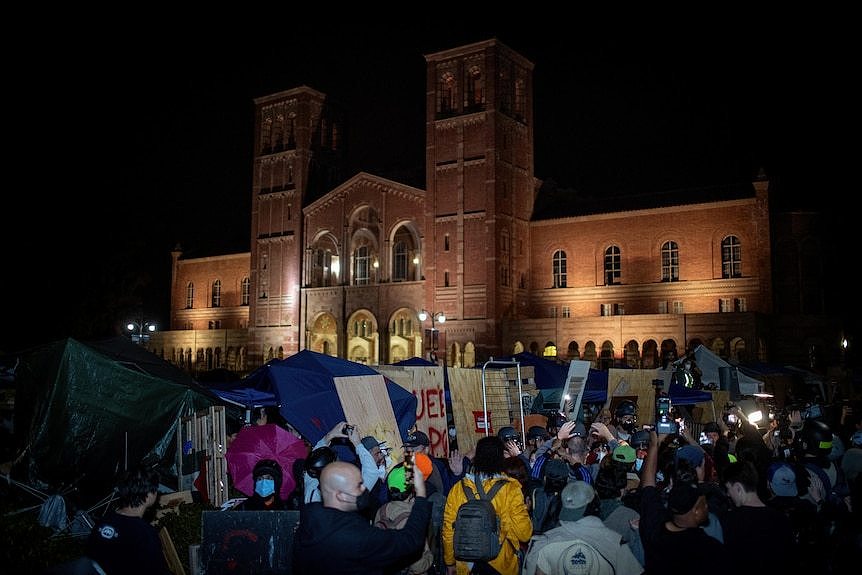 A camp is set up in front of a red brick building. A crowd is gathered in front of it, split by a line of police.
