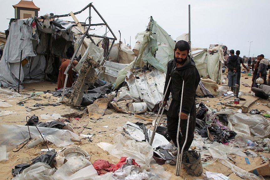 A man leans on crutches surrounded by debris from a damaged tent camp