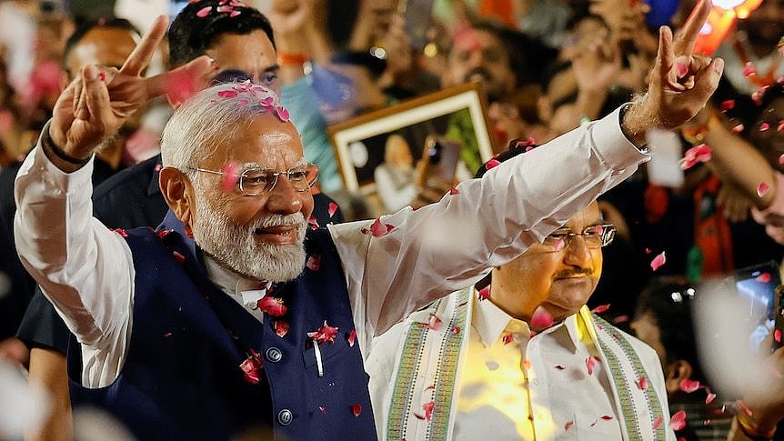 Narendra Modi in a white shirt and blue vest holds his arms in the air making peace signs with his fingers at a rally