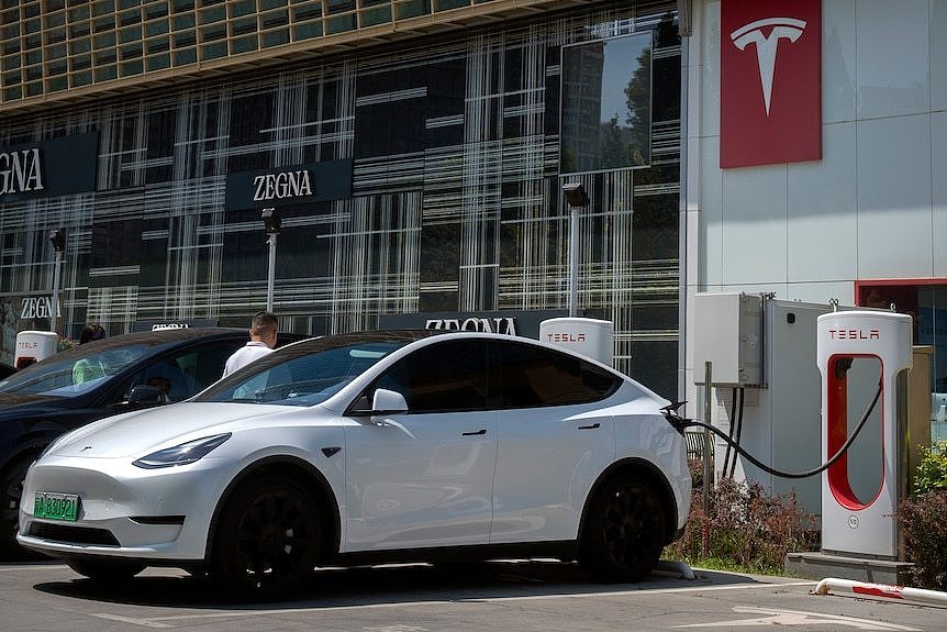 An electric car hooked up to a charger with the Big T Tesla logo on the building behind