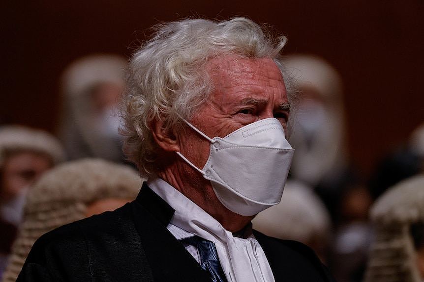 A man with grey curly hair wears a white face mask.