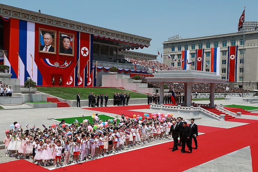 Two men with security guards walk along a red carpet in front of a large building. Nearby is a group of cheering children.