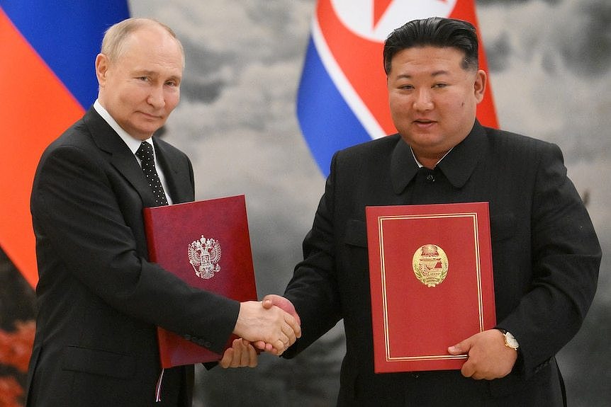 Two men are shaking hands and holding folders while standing in front of country flags.