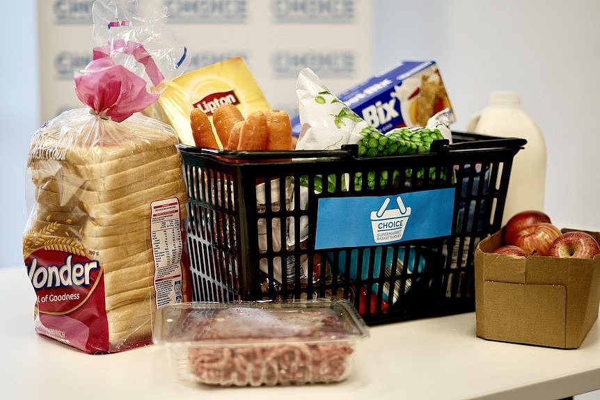 A black shopping basket sitting on a kitchen bench that contains bread, mince, carrots, apples and milk.