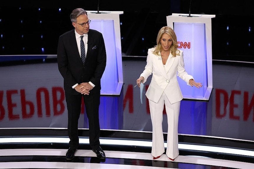 Jake Tapper, in a black suit, and Dana Bash, in a white suit, stand on stage in front of lit-up podiums.