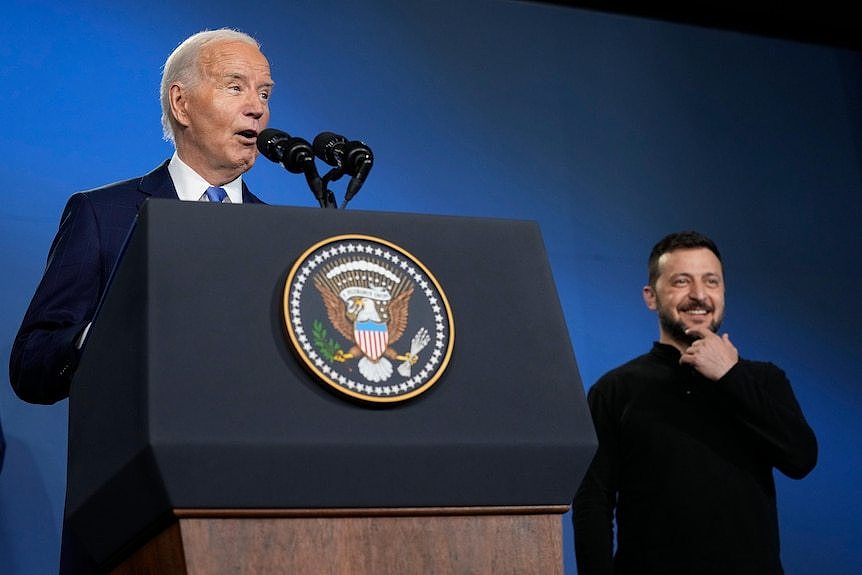 A man standing at a podium wearing a blue suit, in front of a man wearing a black jumper.