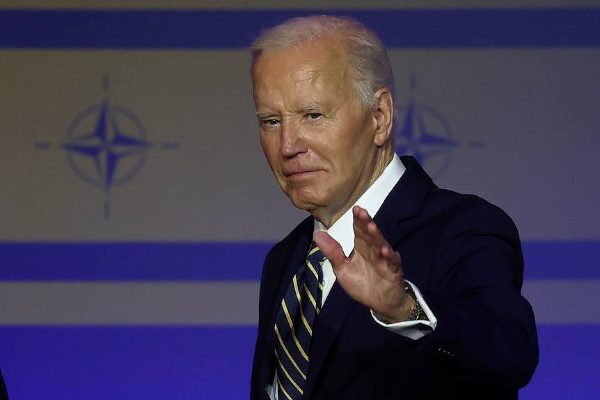 Joe Biden waves at the camera, looking serious, the NATO logo is imprinted on the wall behind him